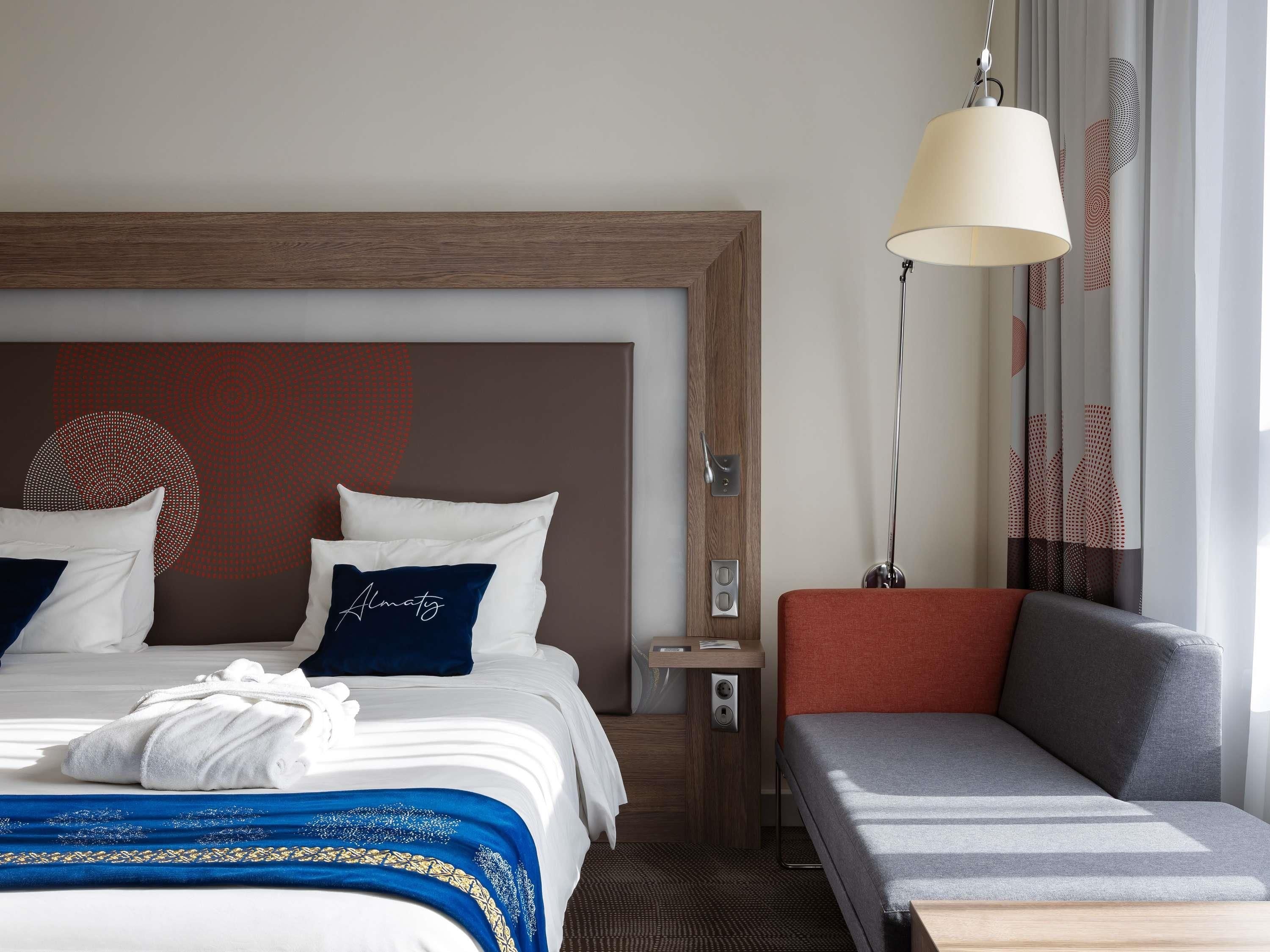 A hotel room with a bed, a bedside table, a lamp, a sofa, and a wall with a geometric pattern.