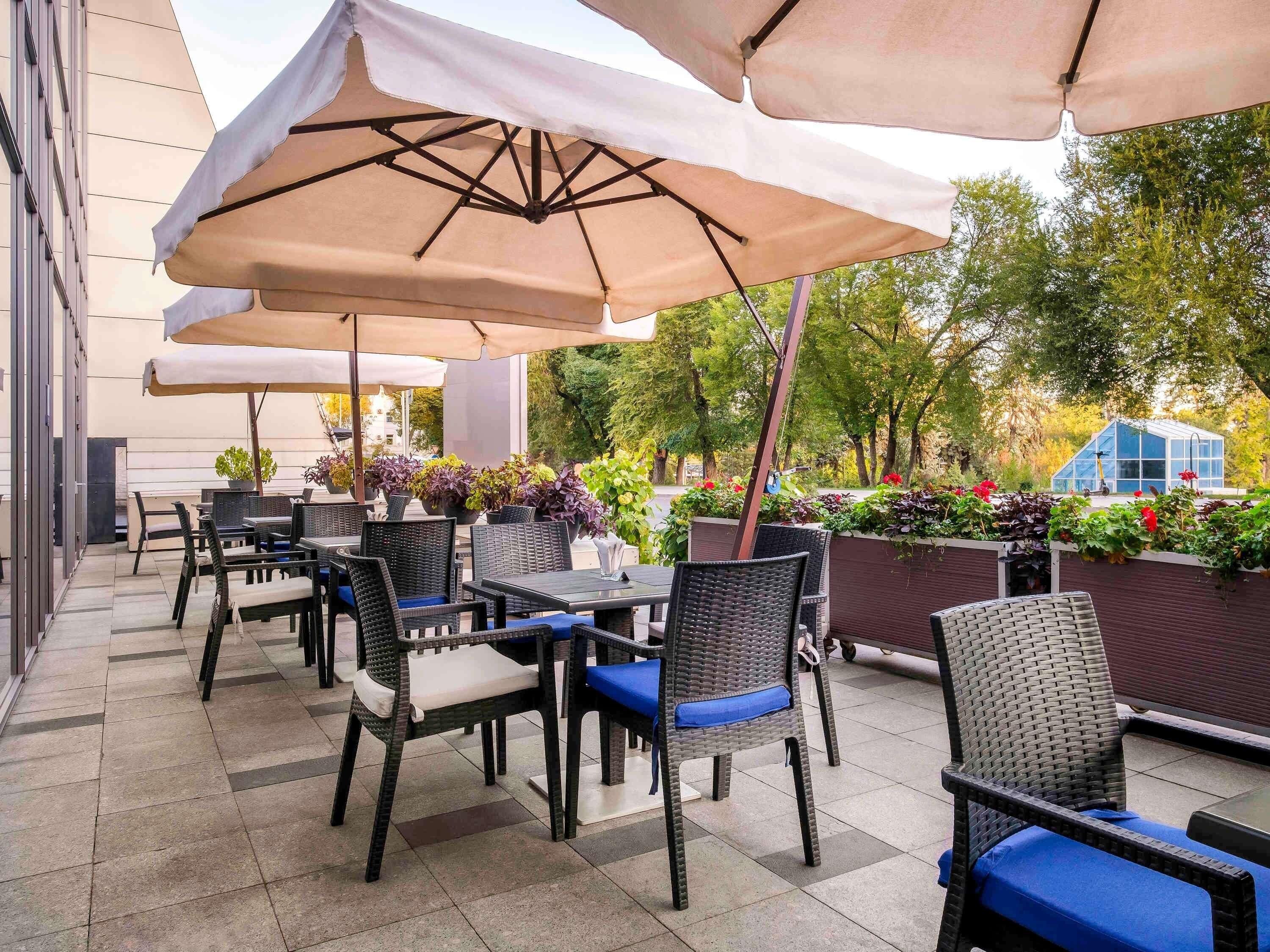 Un espace extérieur aménagé en terrasse, avec des chaises et des tables en osier, protégé par de grands parasols et entouré de plantes en pot et d’une structure de serre en verre.
