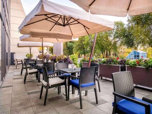 An outdoor patio area with wicker chairs and tables, covered by large umbrellas, and surrounded by potted plants and a glass greenhouse structure. - Novotel Almaty City Center (Almaty)