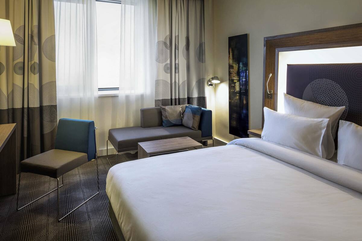 Une chambre d’hôtel avec un grand lit, un bureau, une chaise, un canapé et une vue sur l’extérieur à travers des rideaux légers.