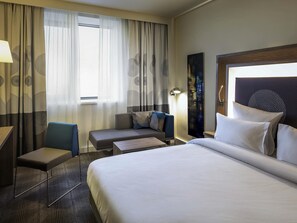 A hotel room with a large bed, a desk, a chair, a sofa, and a view of the outdoors through sheer curtains. - Novotel Almaty City Center (Almaty)