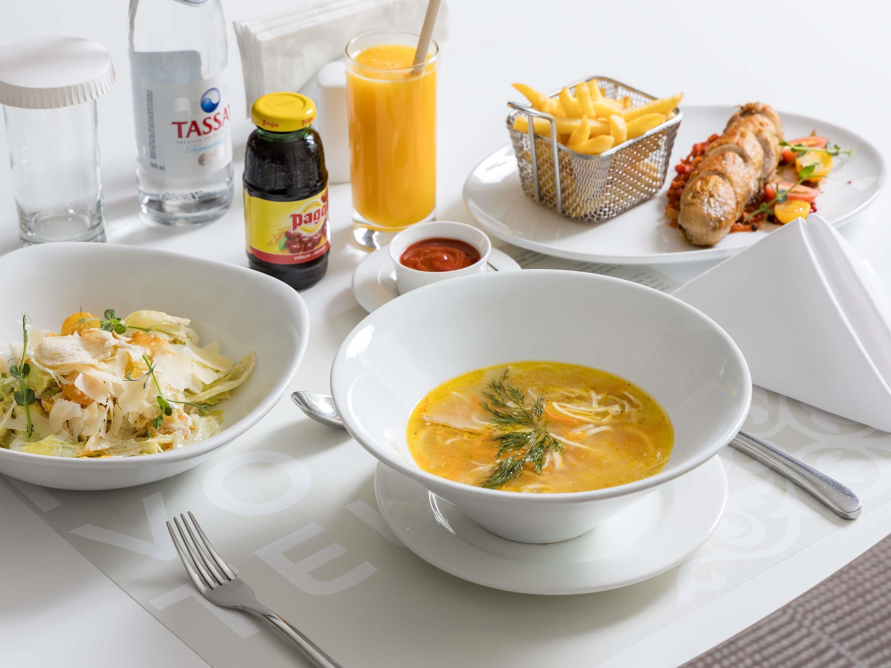 A table with a bowl of soup, a plate of chicken, a basket of fries, a bottle of sauce, a glass of juice, and a bottle of water.
