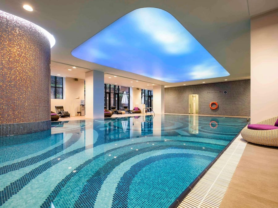 A modern indoor swimming pool with a ceiling skylight, lounge chairs, and a clear view of the sky.