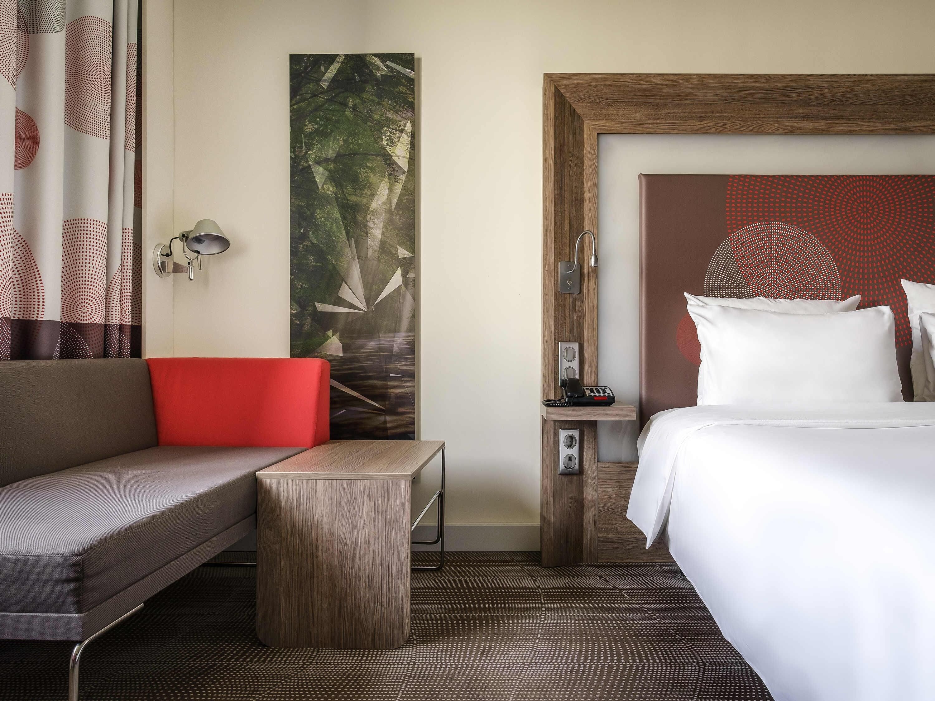 A hotel room with a bed, a sofa, a small table, a wall-mounted phone, and a large abstract painting.
