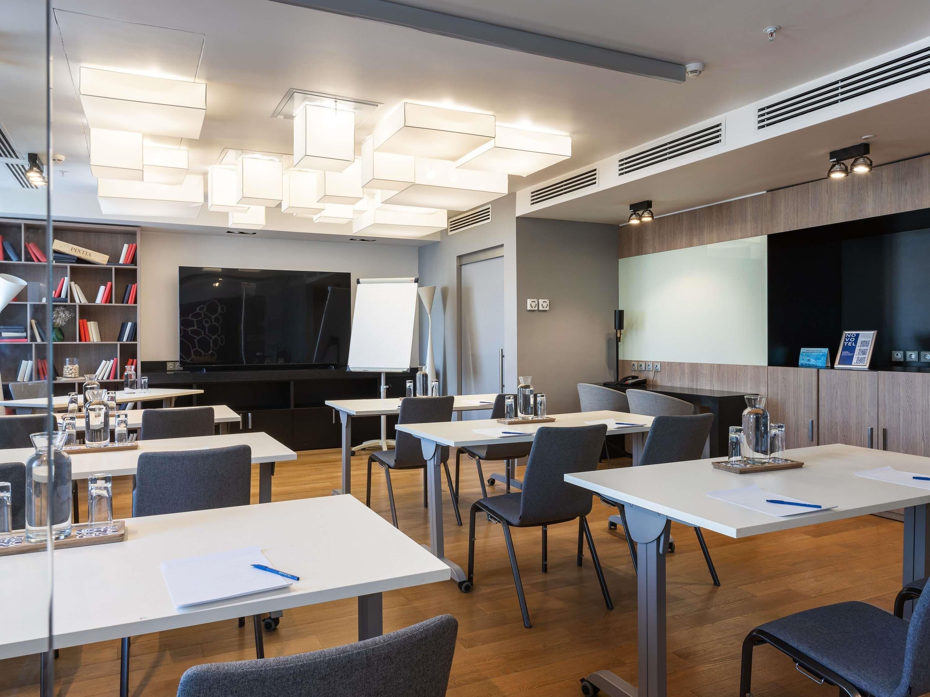 A modern conference room with white tables, grey chairs, a large flat-screen TV, a whiteboard, and a bookshelf with books.