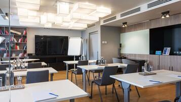 A modern conference room with white tables, grey chairs, a large flat-screen TV, a whiteboard, and a bookshelf with books.