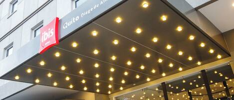Entrada de hotel de diseño moderno, con un llamativo logotipo rojo, zona de acceso cubierta con varias luces y fachada de vidrio que muestra asientos en el interior.