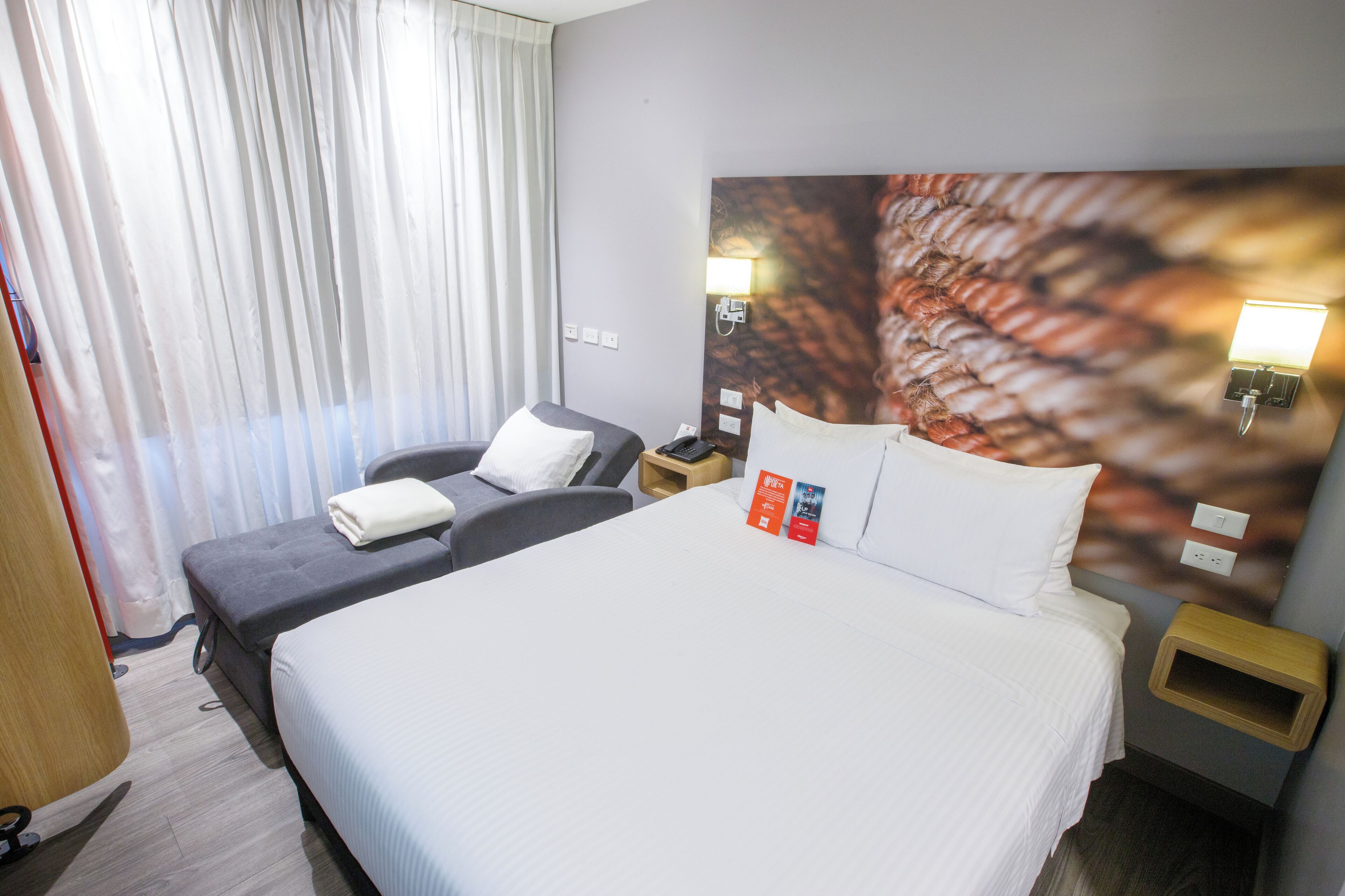 A hotel room with a large bed, a smaller bed, a desk, and a chair. There are two nightstands with lamps and a window with curtains.