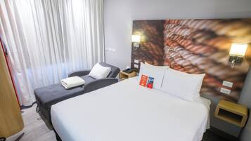 A hotel room with a large bed, a smaller bed, a desk, and a chair. There are two nightstands with lamps and a window with curtains.