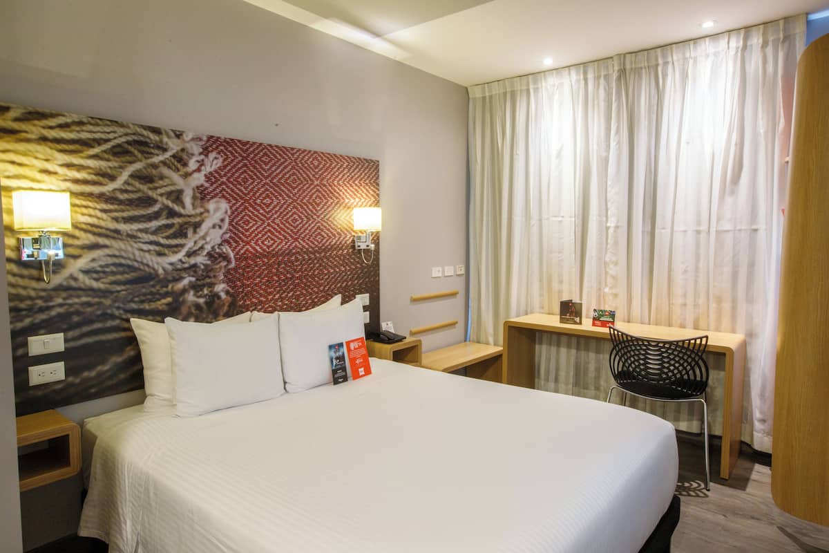 A hotel room with a large bed, a desk, a chair, and a window with curtains.