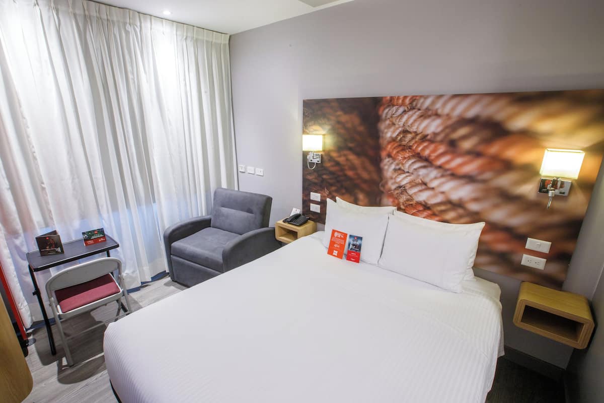 A hotel room with a bed, a desk, a chair, a nightstand, and a wall-mounted lamp.