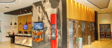 A hotel reception area with a marble counter, a television, a red information kiosk, and a wall with a cityscape mural.