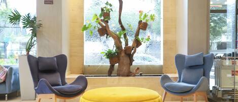 A lobby with two grey armchairs, a yellow ottoman, and a large window with a tree sculpture.