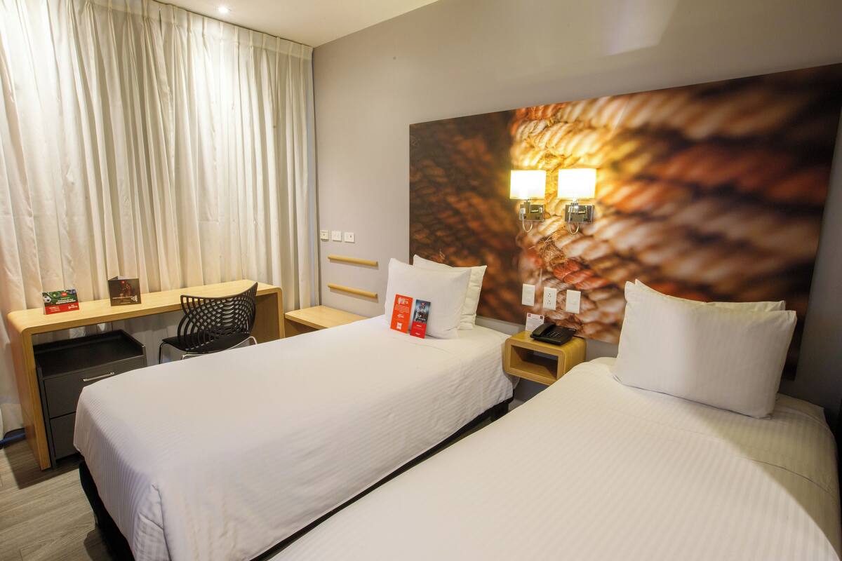 A hotel room with two beds, a desk, a chair, and a large wall art piece.