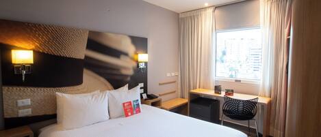A hotel room with a large bed, bedside tables, a desk, a chair, and a window with curtains.