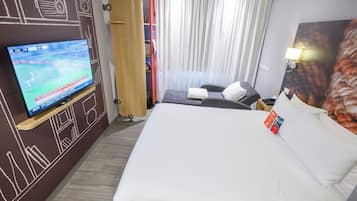 Habitación de hotel con una cama grande, un televisor en la pared, un escritorio con lámpara y una ventana con cortinas.