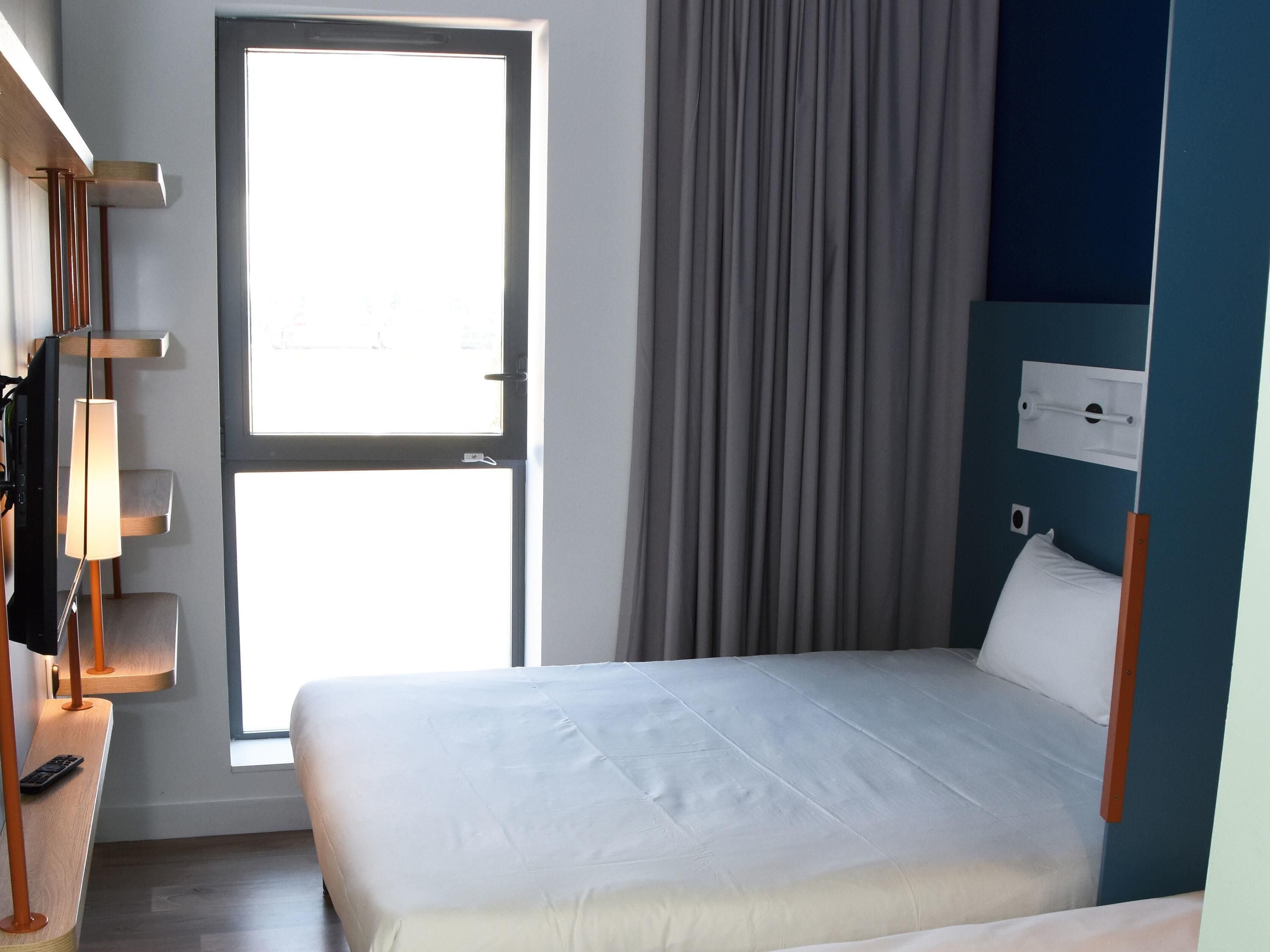Una habitació d'hotel amb un llit, un escriptori amb una làmpada, una finestra amb cortines i un panell de control fixat a la paret.