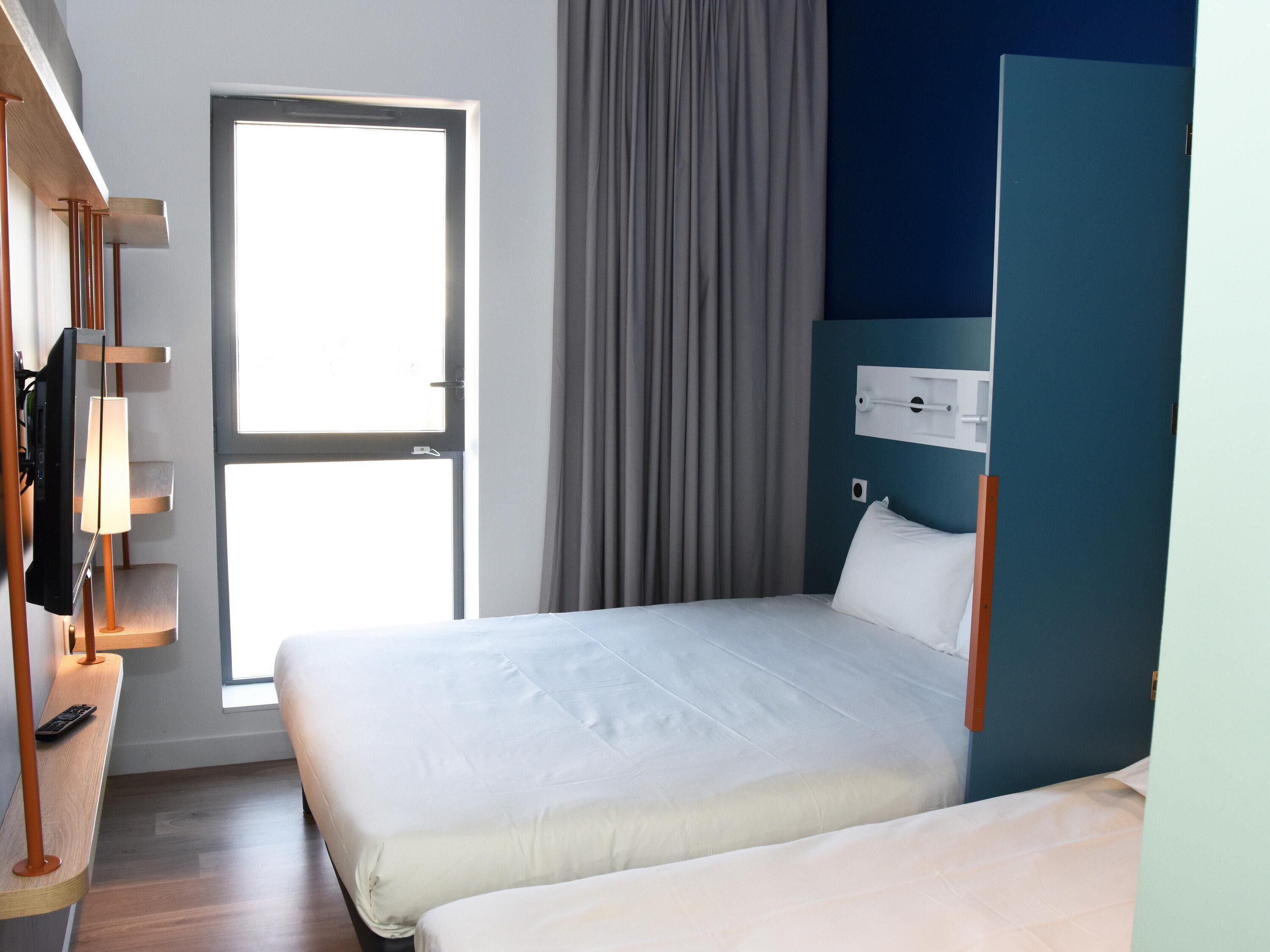 A hotel room with a bed, a TV, a desk with a lamp, and a window with curtains.