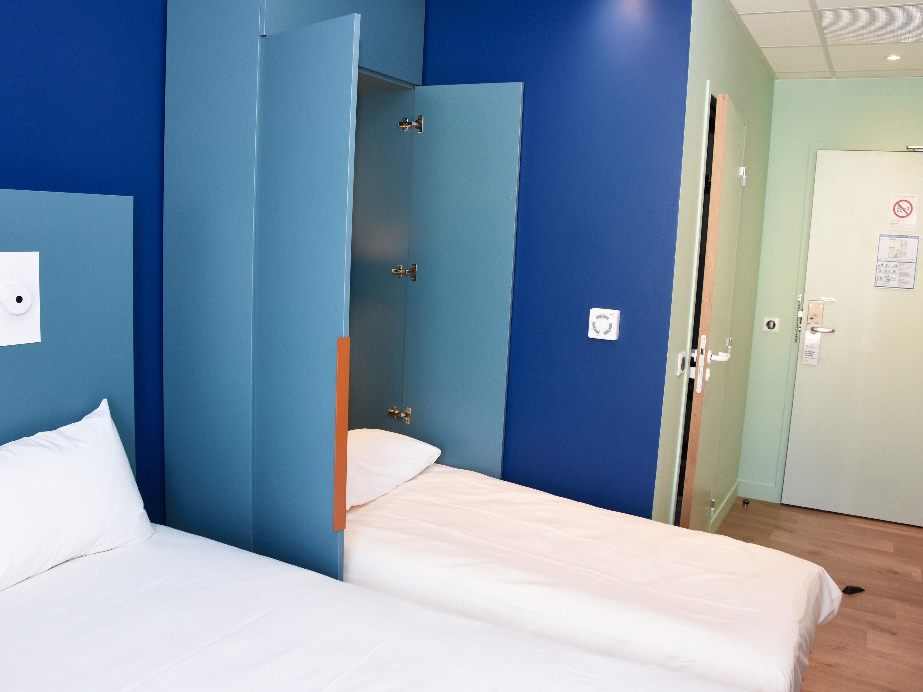 A hotel room with two beds, a wardrobe, a door with a sign, and a light switch on the wall.