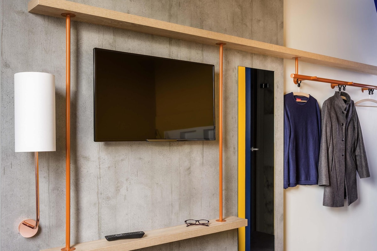 Een moderne kamer met een wandgemonteerde televisie, een houten plank met een afstandsbediening, en een kledingrek met twee kledingstukken.