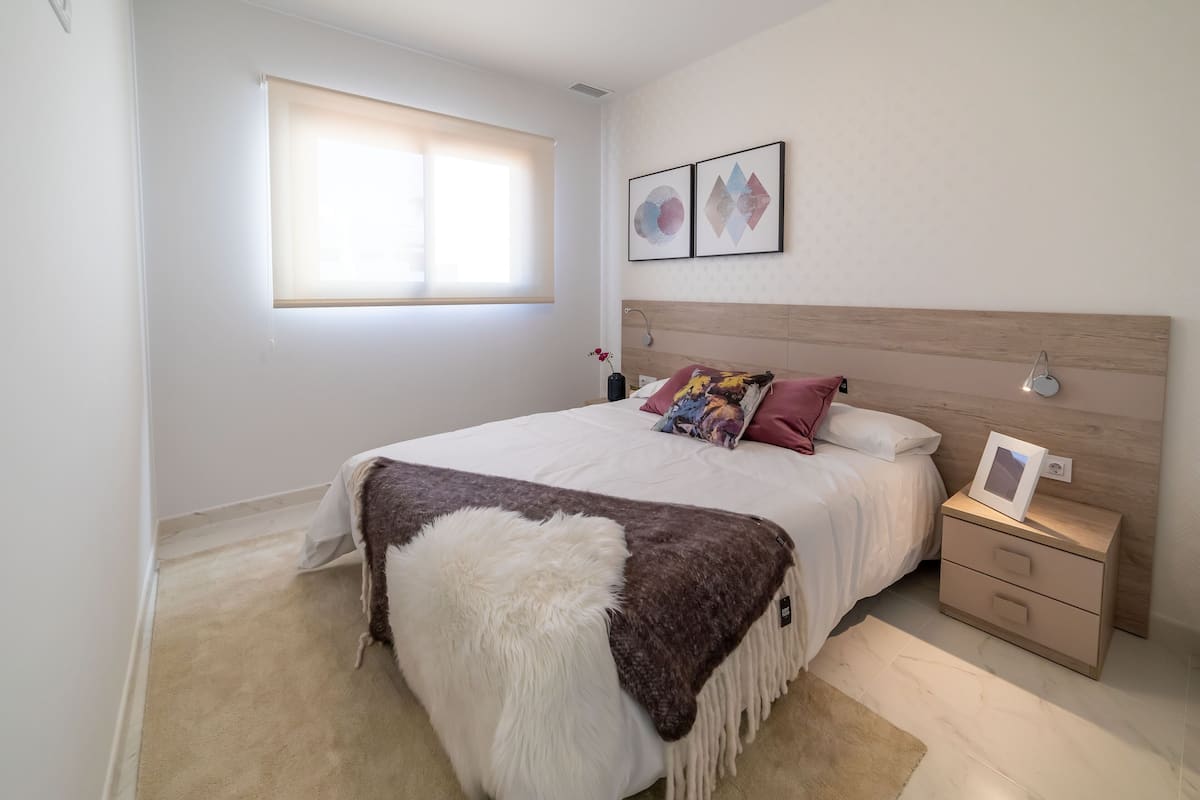 A bedroom with a bed, a nightstand, a window with blinds, and two wall-mounted artworks.