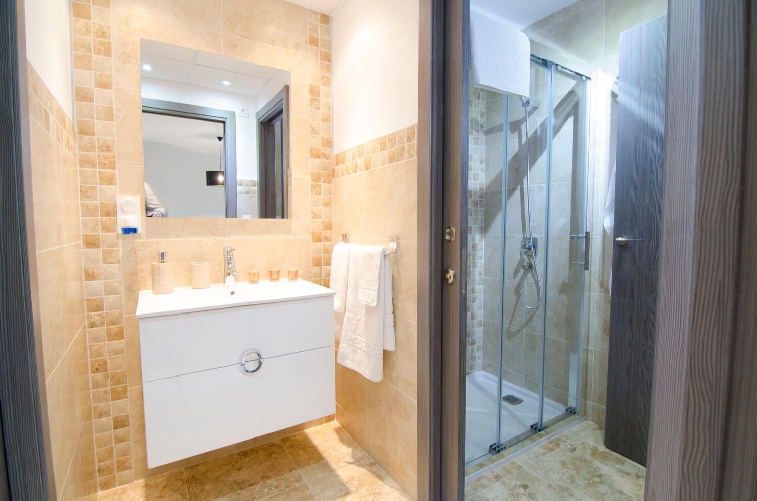 Un baño moderno con ducha envuelta en vidrio, tocador blanco con espejo y paredes de baldosas color beige.