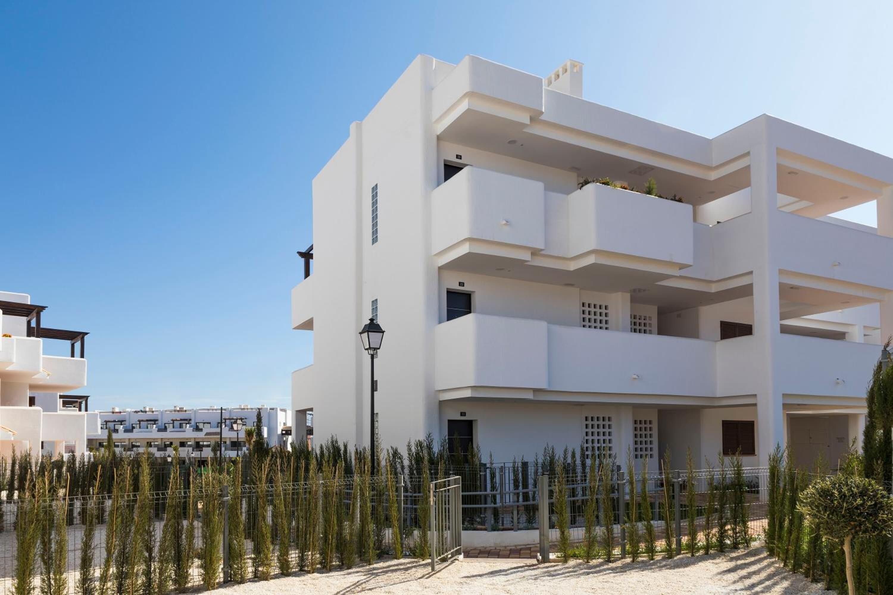 A modern multi-story building with balconies, a gated entrance, and a clear sky.