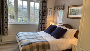 A neatly made bed with a striped blanket and two pillows, a bedside table with a lamp, and a window with curtains. - Beck Allans Guesthouse (Ambleside)