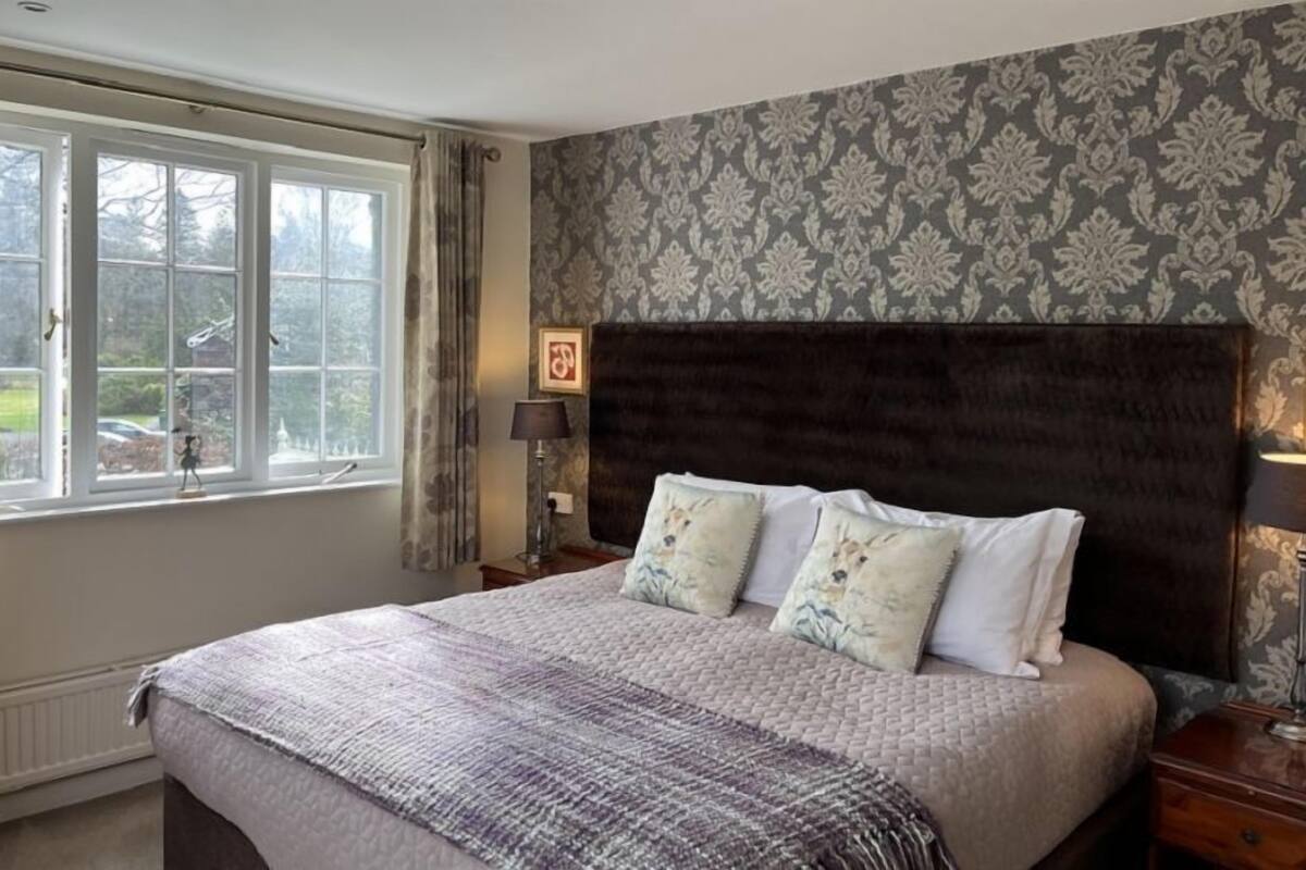 A bedroom with a large bed, a window with curtains, a nightstand with a lamp, and a patterned wallpaper.