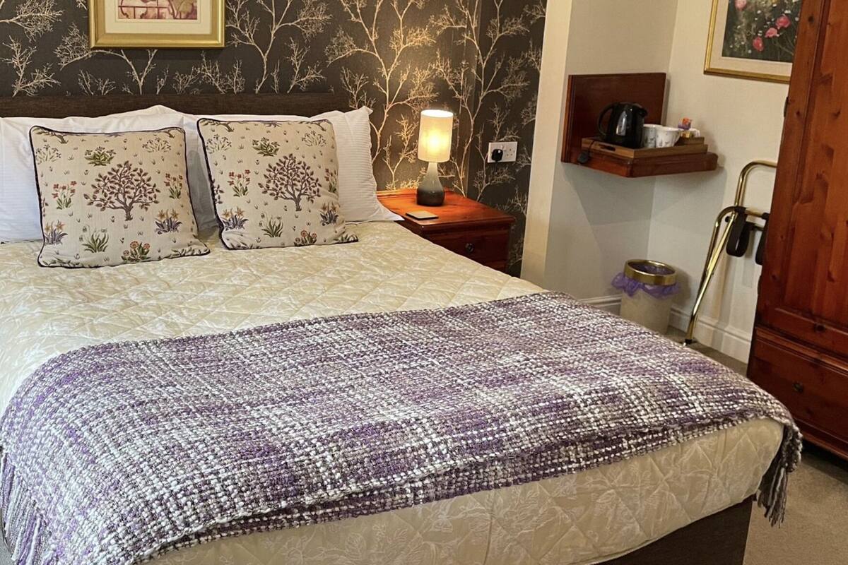 A neatly made bed with floral-patterned pillows and a purple and white blanket. There are two bedside tables with lamps and a small wooden nightstand with drawers. The room has dark wallpaper with a tree pattern and a wooden wardrobe.