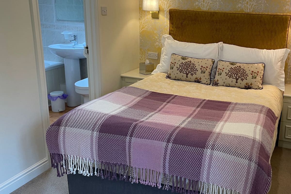 A neatly made bed with a checkered blanket, two pillows with floral patterns, and a bedside table with a vase of dried flowers.