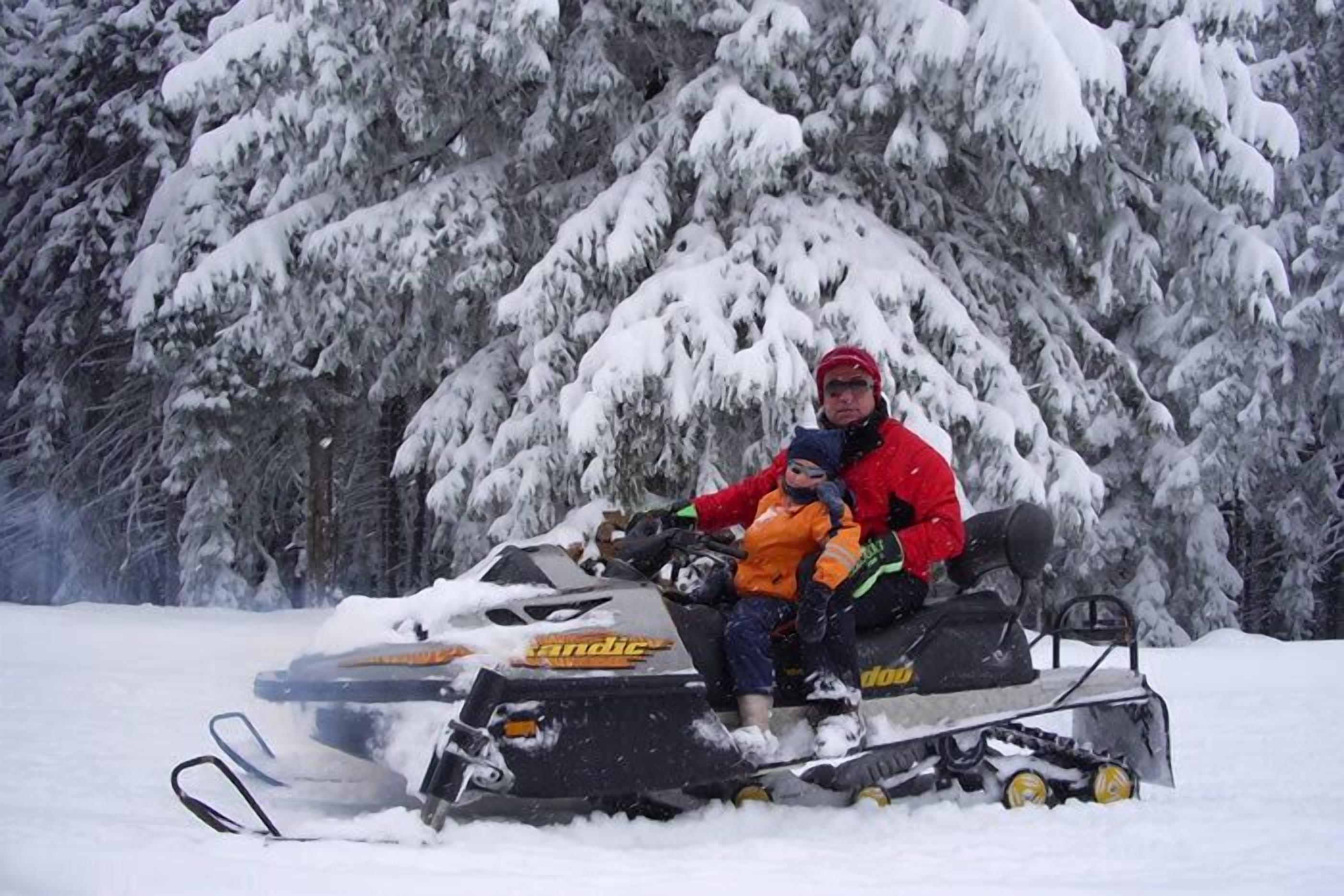 Een sneeuwscooter met een kind vooraan, voor een besneeuwd bos.