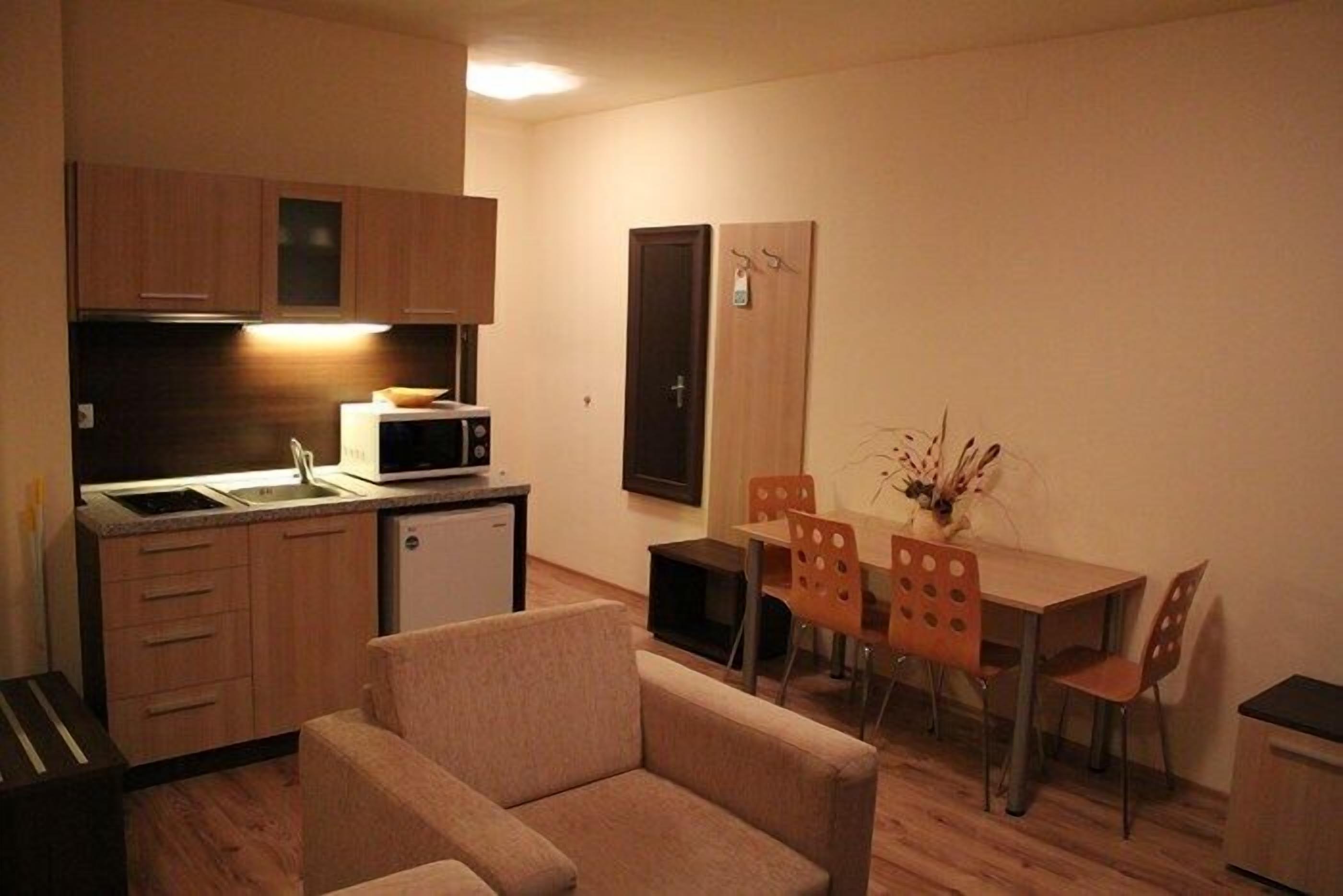 A compact kitchen with a microwave, a small refrigerator, and wooden cabinets. There is a dining table with four chairs and a sofa in the living area.