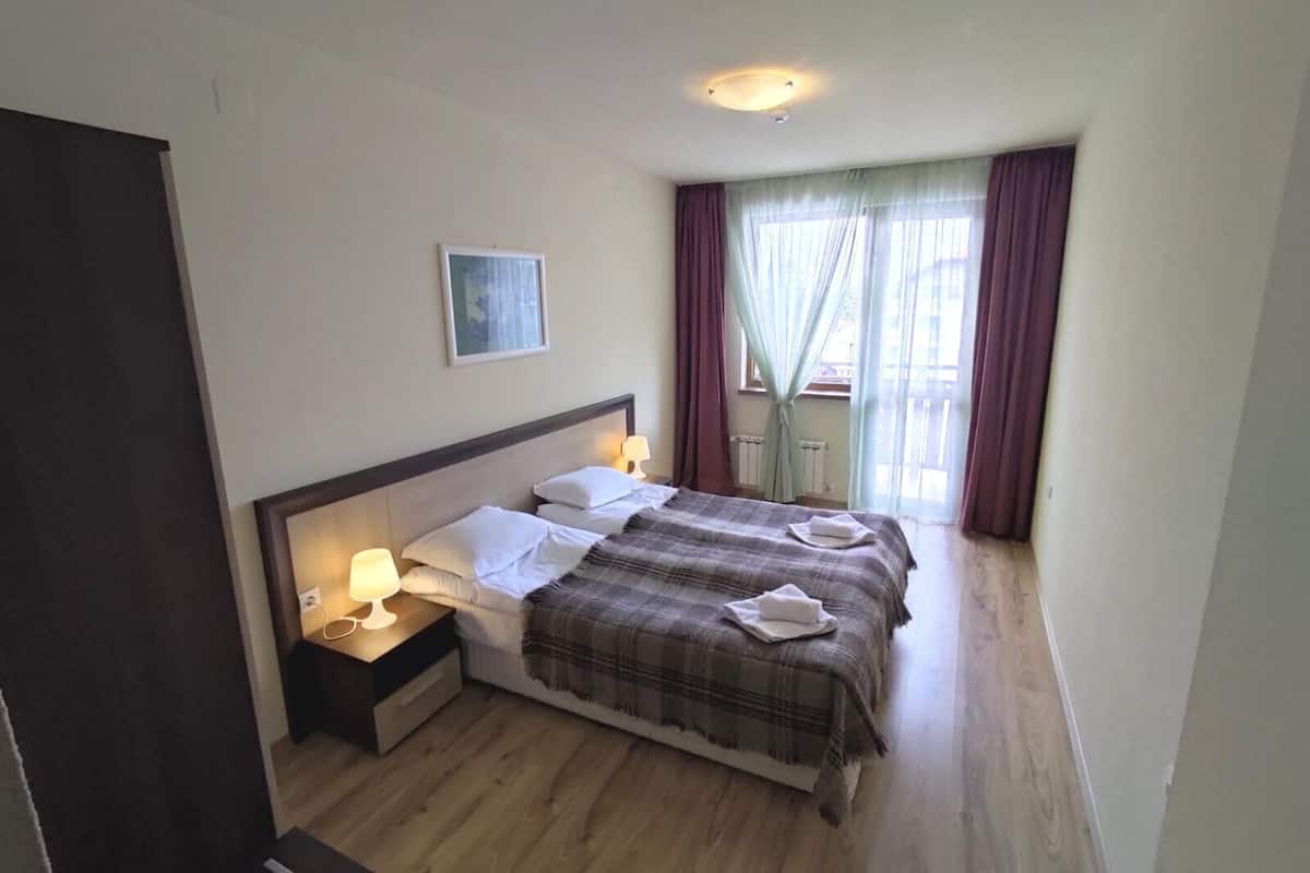 A bedroom with a bed, bedside lamps, a framed picture on the wall, and a window with curtains.