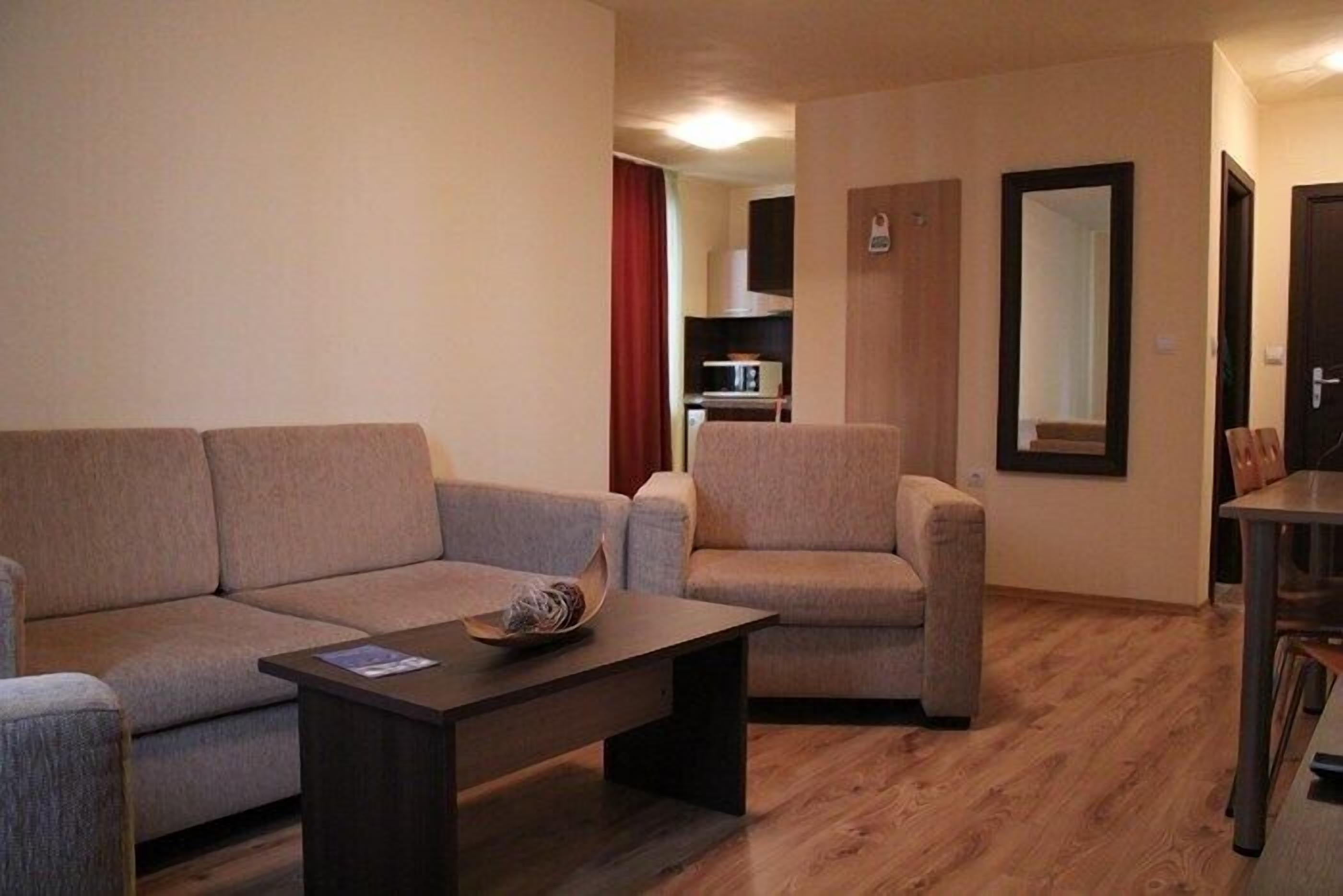 A living room with a sofa, armchair, and coffee table. There is a door leading to a kitchen area.