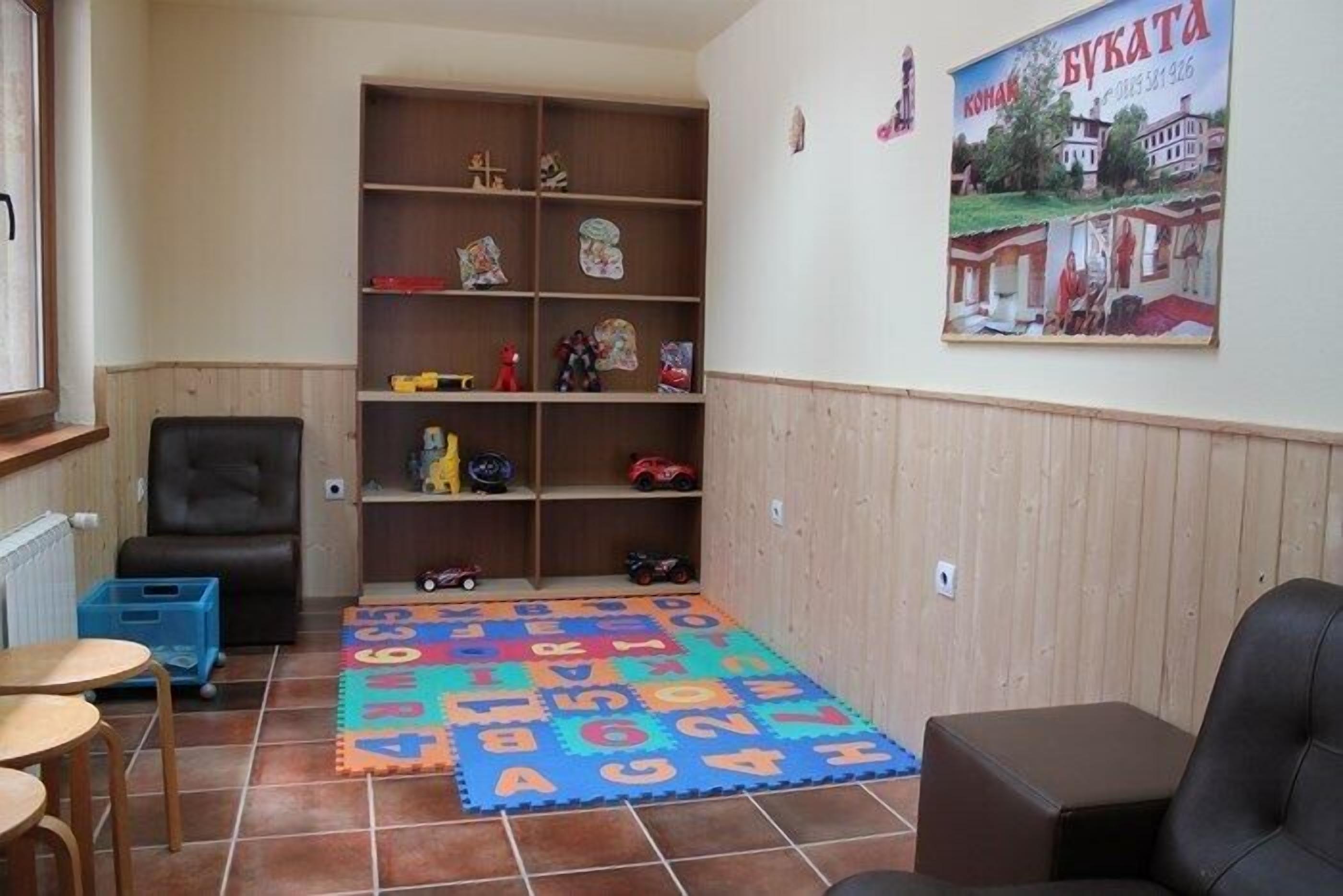 Een kamer met een speelruimte, voorzien van een kleurrijke letterfoam-mat, een boekenkast met speelgoed en een poster aan de muur.