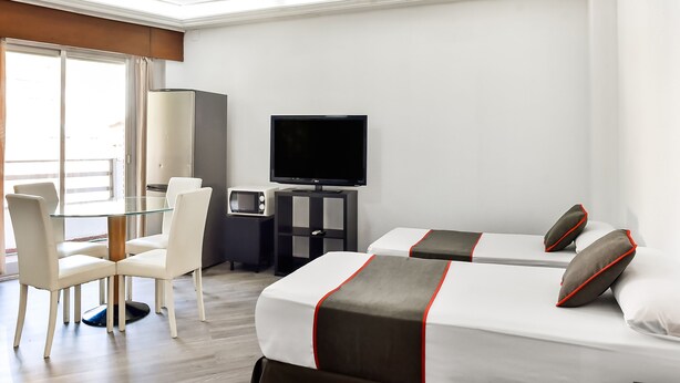 Habitación de hotel con dos camas, un escritorio con computadora, televisor y zona de estar con sillones.