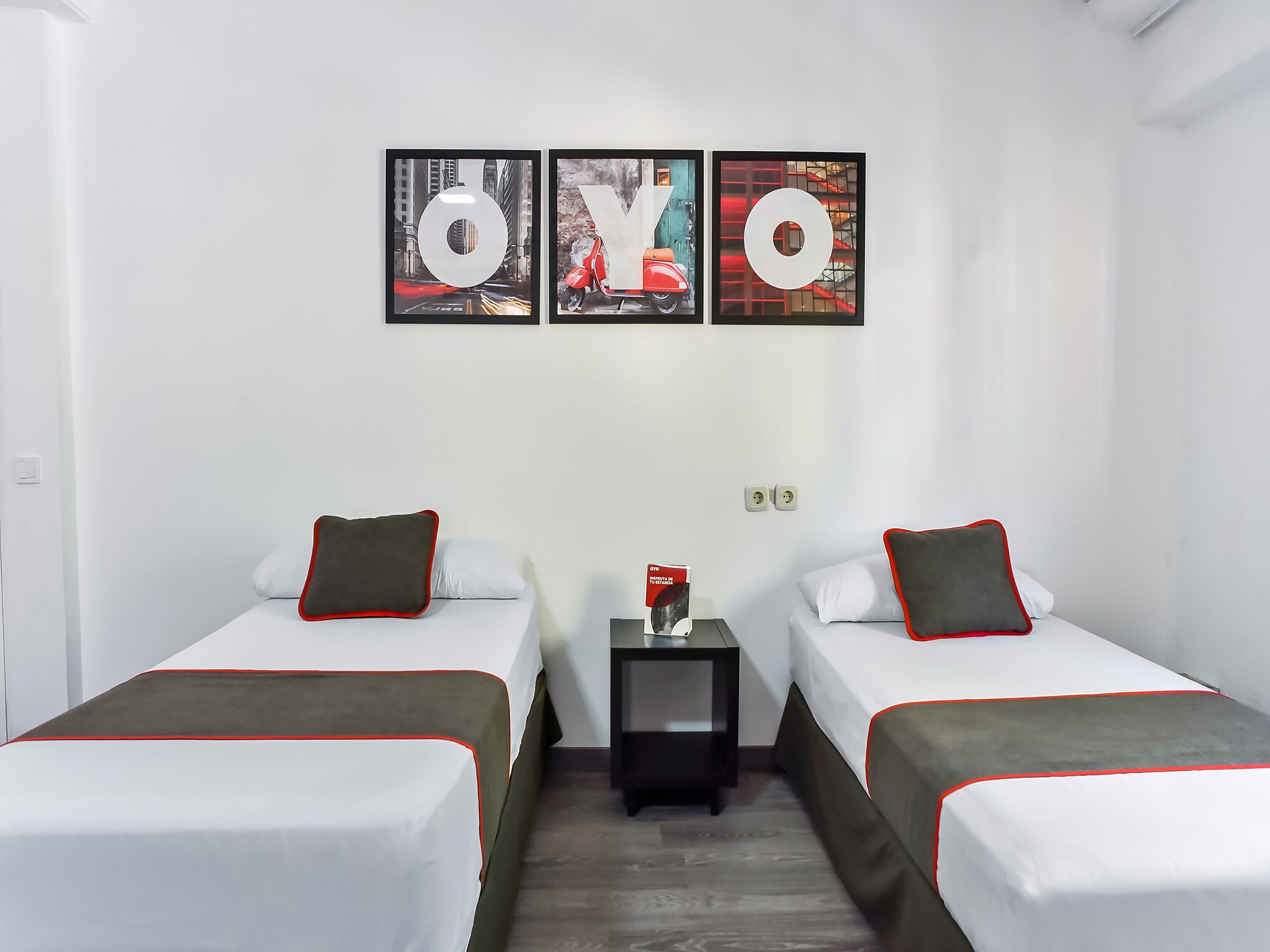 Habitación de hotel con dos camas, cada una con ropa blanca y detalles rojos, una mesita de noche con un libro y tres cuadros en la pared que deletrean "OYO".