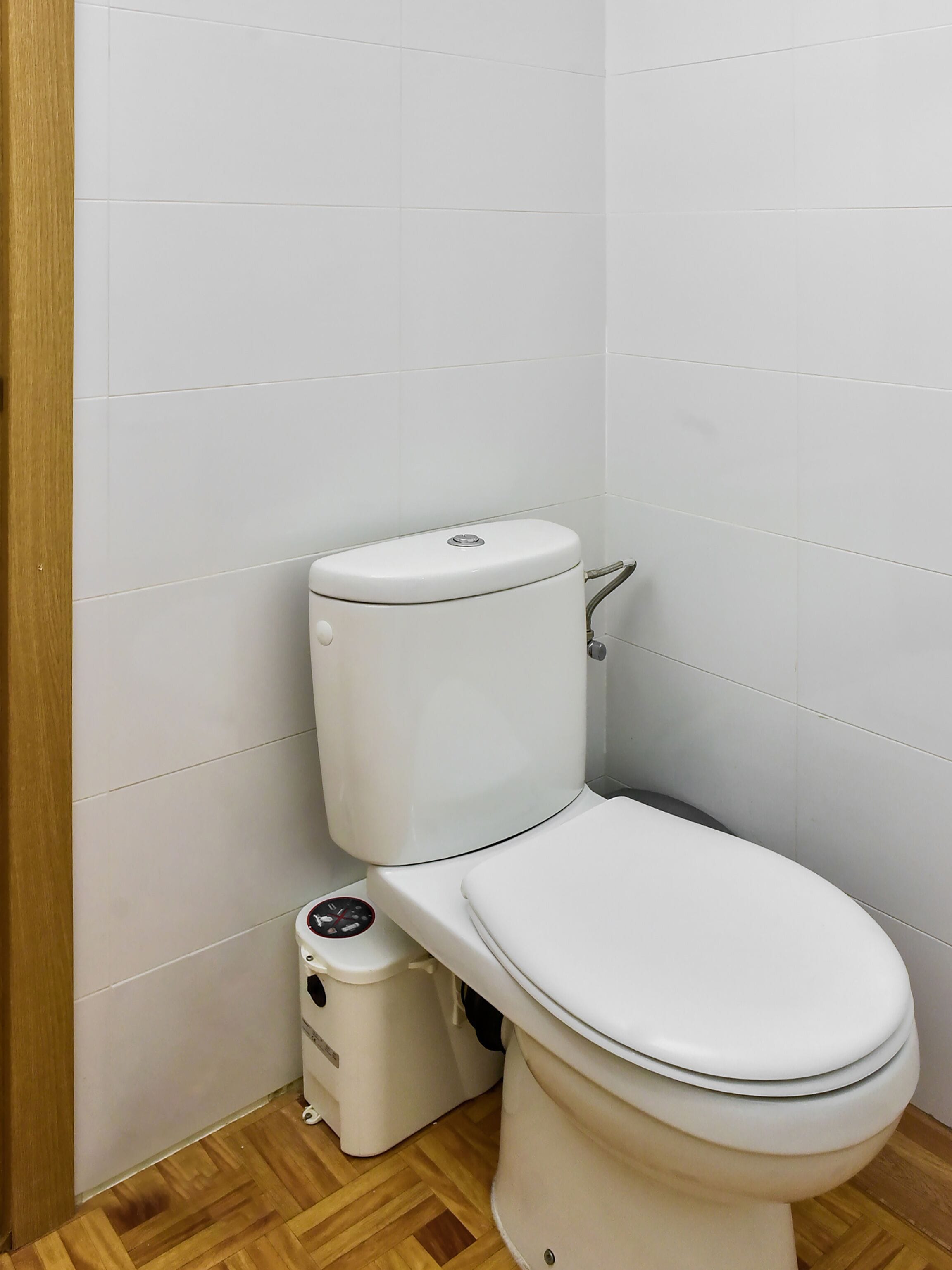 A toilet with a flush handle and a bidet attachment in a bathroom with white tiled walls and wooden flooring.