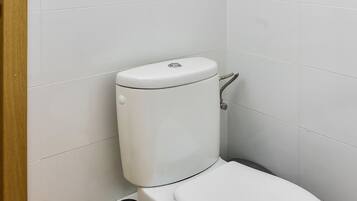 A toilet with a flush handle and a bidet attachment in a bathroom with white tiled walls and wooden flooring.
