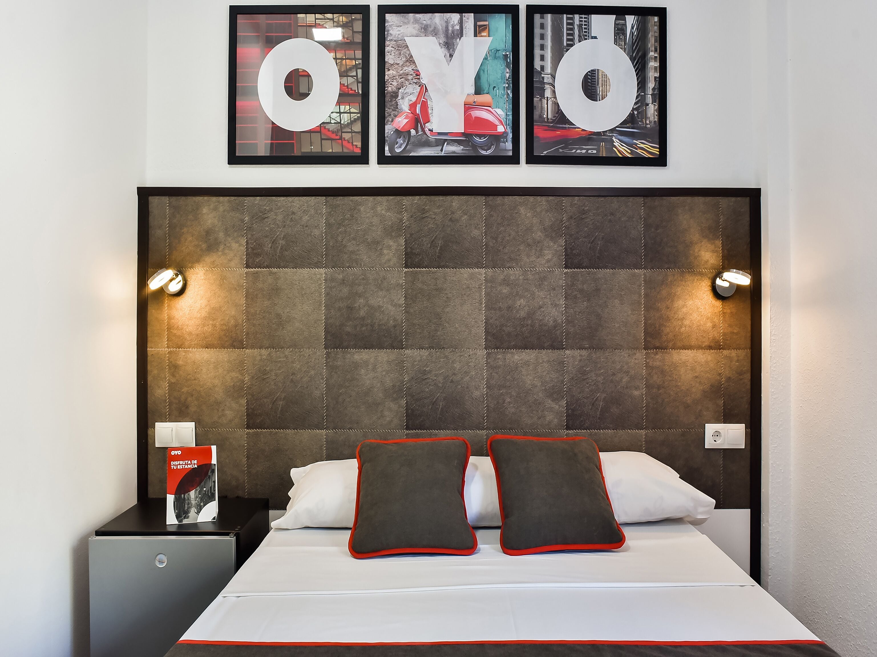 Ein Hotelzimmer mit einem großen Bett, zwei Kissen, einem Nachttisch mit einer Zeitschrift und drei gerahmten Bildern an der Wand über dem Bett.
