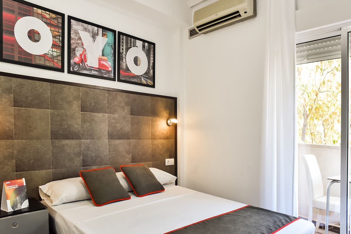 Una habitación de hotel con una cama, una mesita de noche, una obra de arte enmarcada sobre la cama y una ventana con vistas a árboles.
