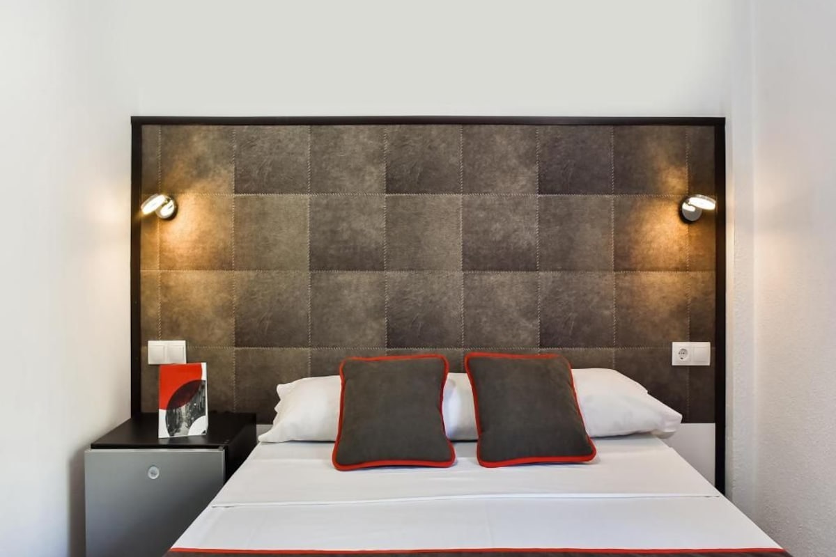 Habitación de hotel con una cama grande, dos lámparas de noche, una mesita de noche con un libro y un cabecero moderno.