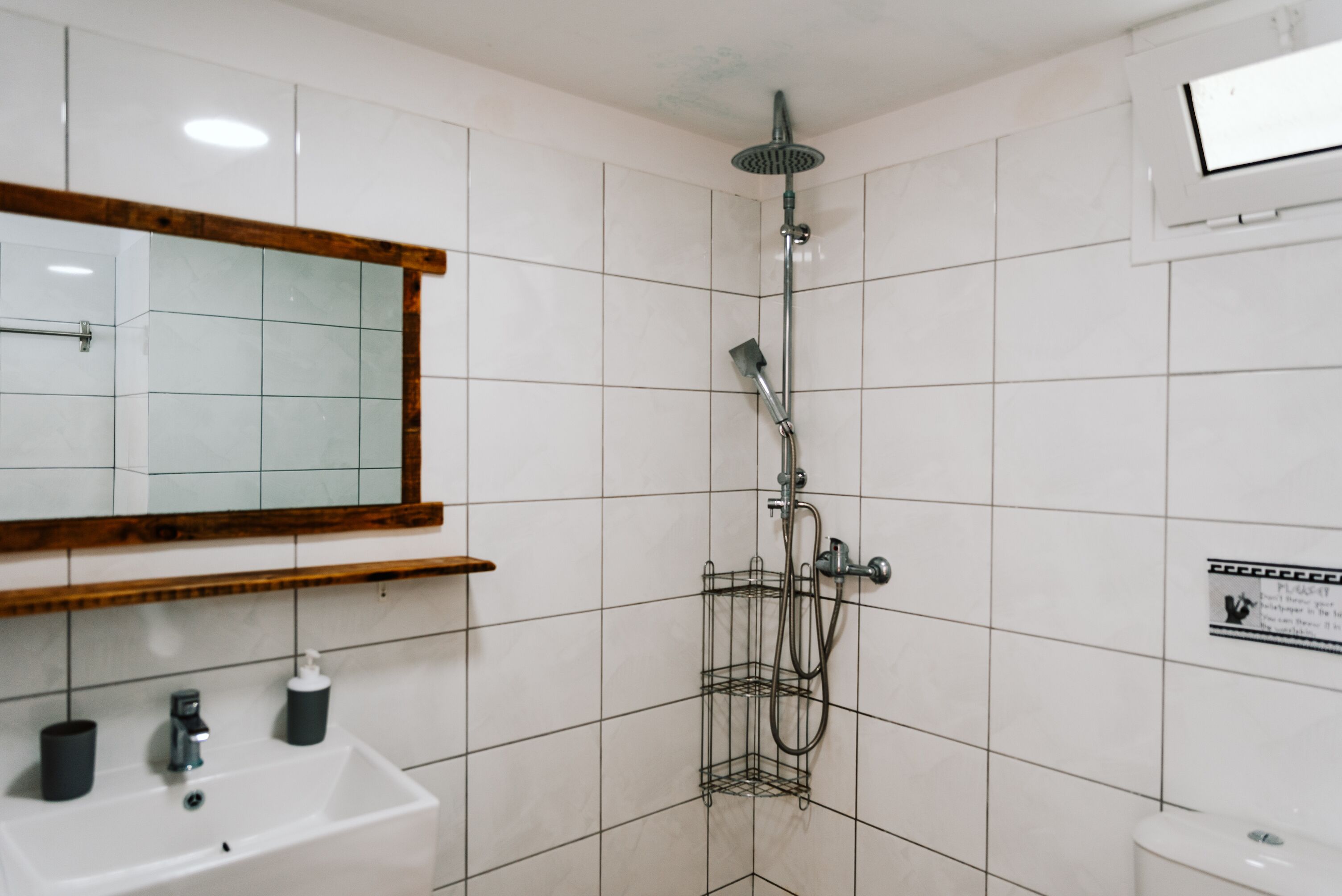 A bathroom with white tiles, a wooden-framed mirror, a white sink, a shower with a handheld showerhead, and a toilet.