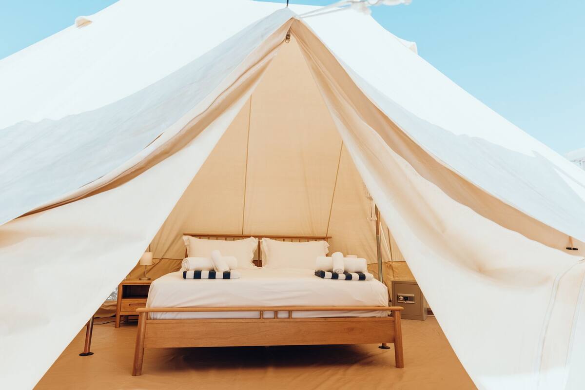 Una cama dentro de una tienda de campaña con estructura de madera y ropa de cama blanca, rodeada de paredes de tela blanca.