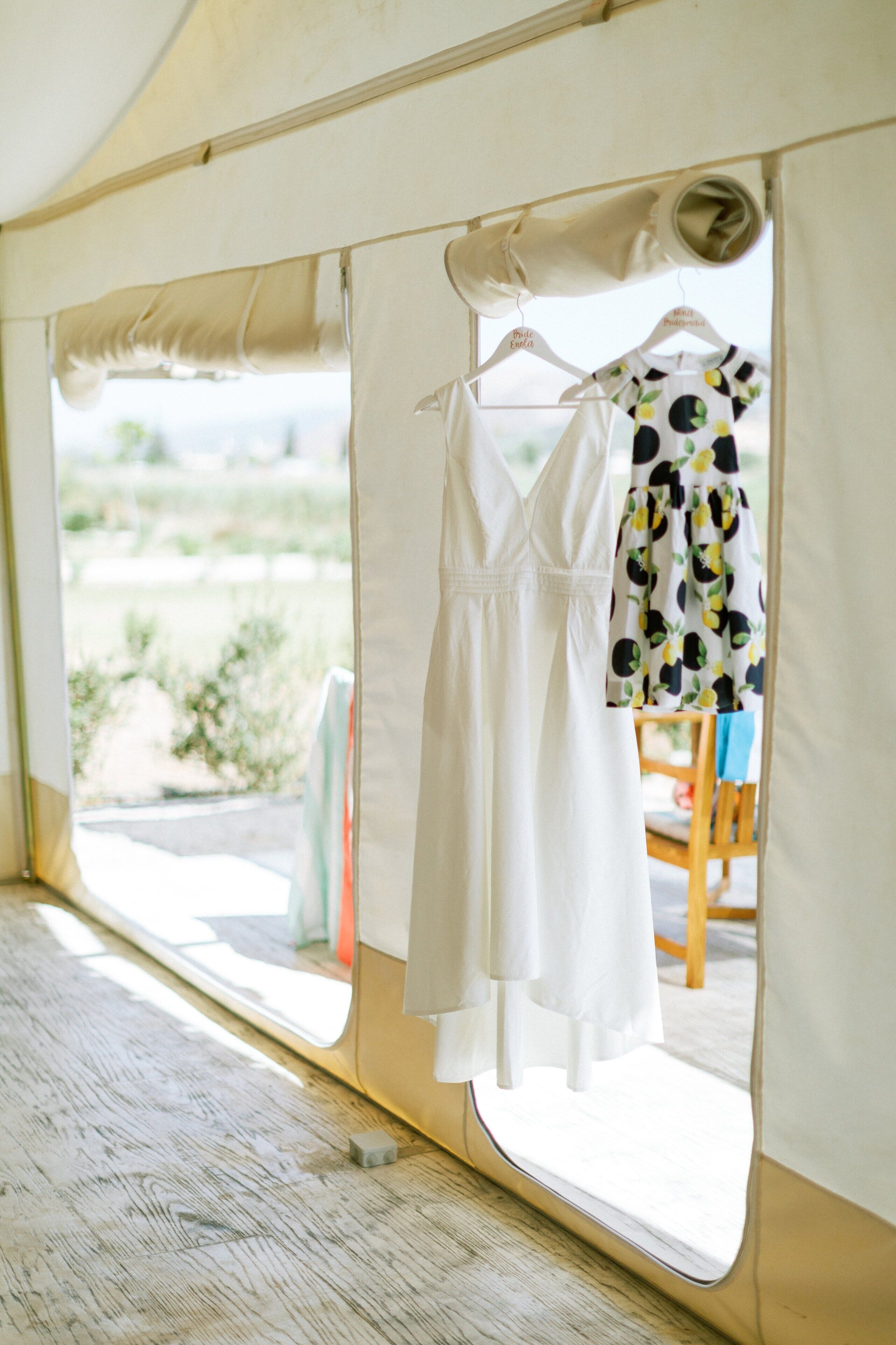A tent interior with a white dress hanging on a hanger and another dress with a fruit pattern on a chair.