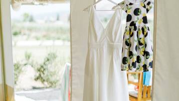 A tent interior with a white dress hanging on a hanger and another dress with a fruit pattern on a chair.