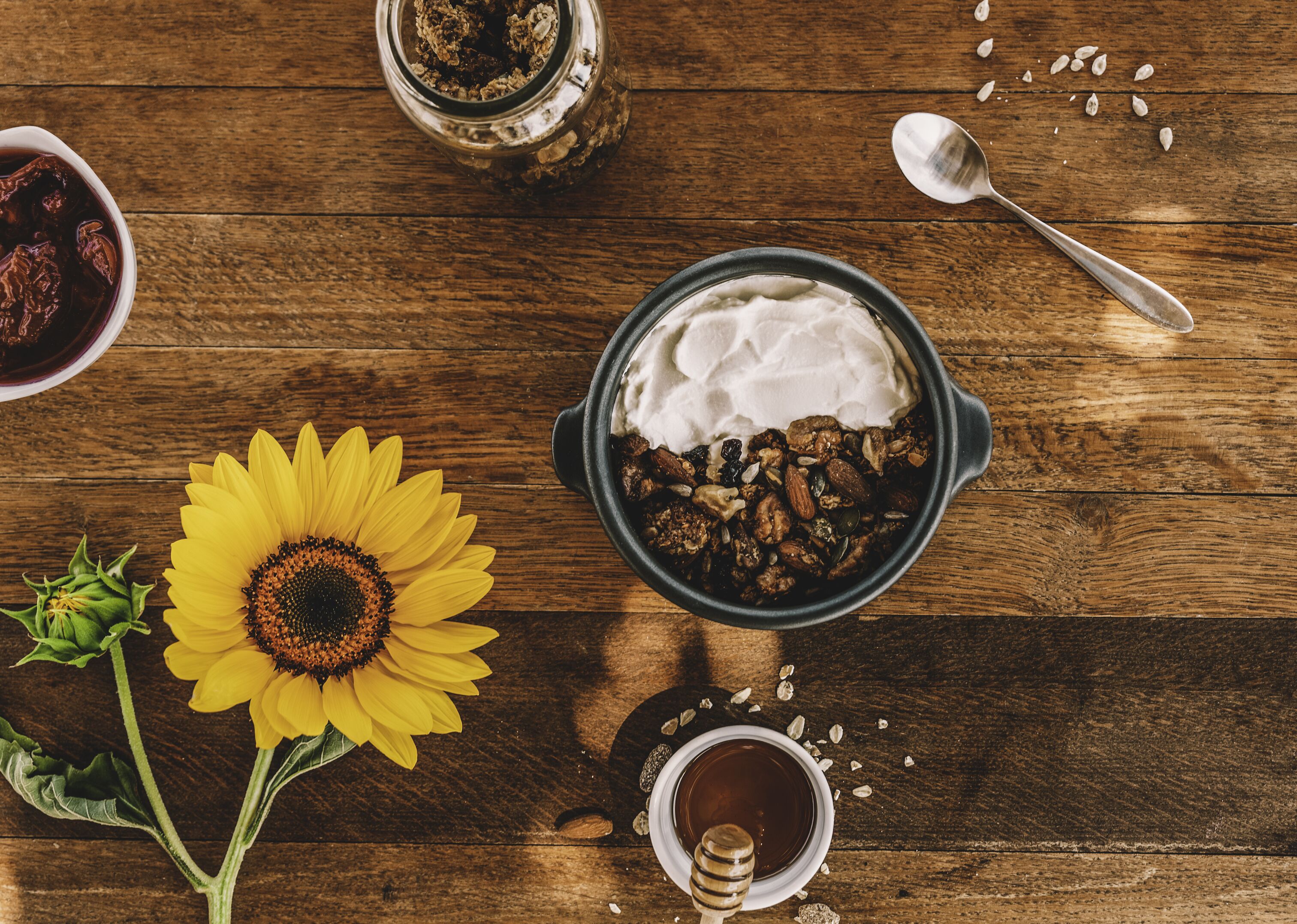 A wooden table with a bowl of granola, a bowl of yogurt, a jar of jam, a honey dipper, and a sunflower.