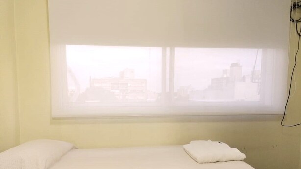 Ein Einzelbett mit weißer Bettdecke und Kissen, ein Fenster mit Blick auf Gebäude und eine wandmontierte Steckdose.