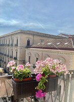 Un balcone con vasi di gerani e vista su un edificio storico con finestre ad arco e tetto in tegole.